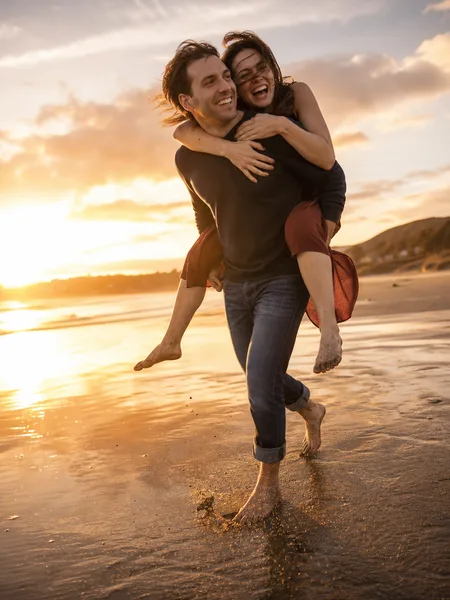 Piggyback ride on a grey sand beach at golden hour, wearing burgundy dress and navy sweater, engagement photos.
