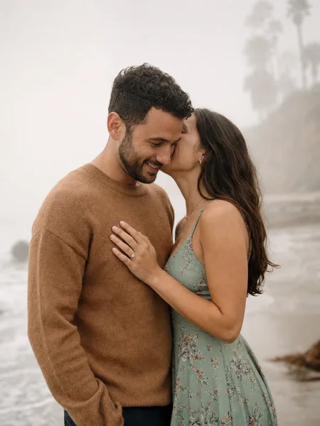 Engagement photo of a couple on a foggy beach, the woman whispering into the man's ear, wearing a floral midi dress, soft misty background.
