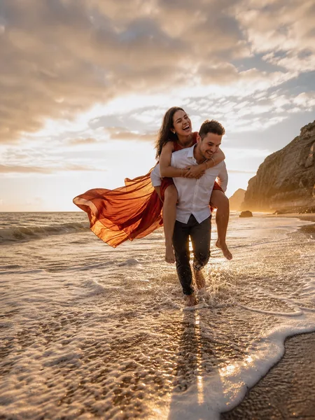 Groom giving bride a piggyback ride on a grey sand beach at golden hour, her burgundy dress flying, romantic engagement photos.