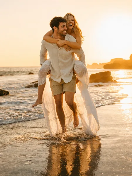 Wide shot of couple giving piggyback ride in shallow ocean water at golden hour, white dress flowing, engagement photos.