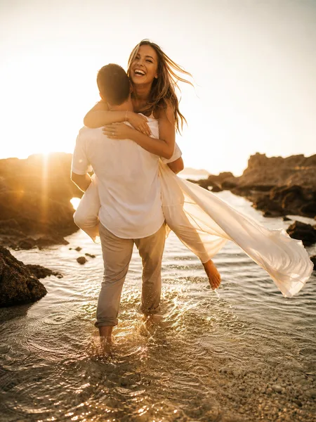 Groom carrying bride piggyback in a tidepool at golden hour, white dress flowing, splashing water, warm backlight engagement photos.