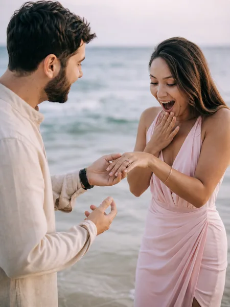Close-up of a couple on the beach at morning, he reveals a ring as she reacts with surprise, pastel pink dress, engagement photos.