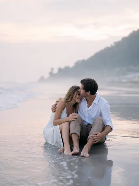 Couple sitting on wet sand at water's edge at sunrise, misty beach, casual sundress, engagement photos.
