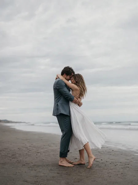 A couple slow dancing barefoot on a grey sand beach under an overcast sky, editorial engagement photos.