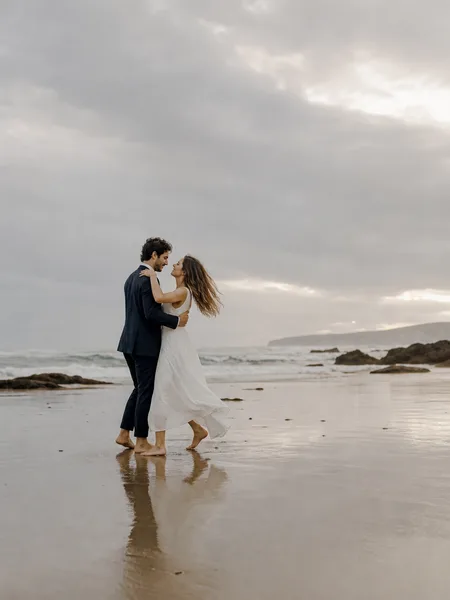 Couple slow dancing barefoot on an overcast beach, white dress flowing, editorial engagement photos.