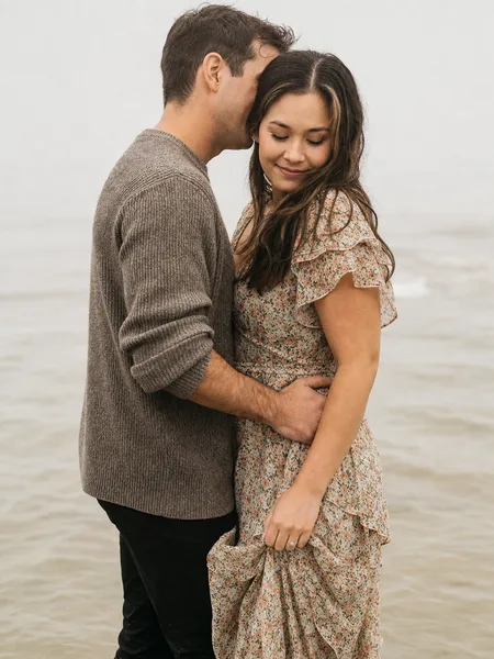 Tight portrait of couple whispering in shallow water on foggy beach, floral dress and sweater, engagement photos.
