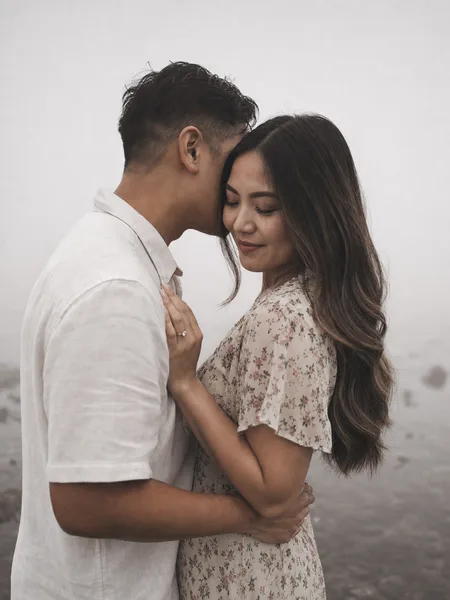 Close-up of couple whispering in thick fog by a tidepool, floral midi dress, moody light, intimate engagement photos