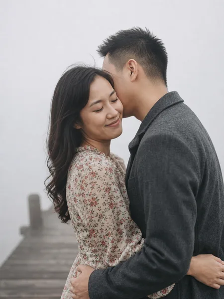 Close-up of man whispering in woman's ear on foggy wooden pier, floral dress, intimate engagement photos.