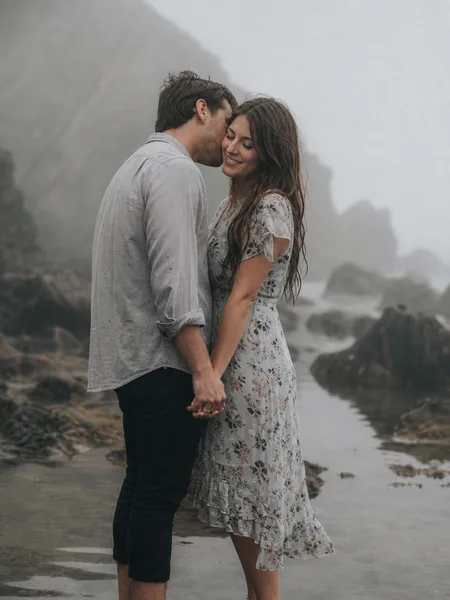 Close-up of a couple whispering in a foggy tidepool, floral dress, shallow water, intimate and moody engagement photos.
