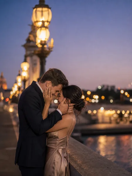 Couple in formal attire touching foreheads on an old bridge, city lights glowing in the dusk, engagement photos