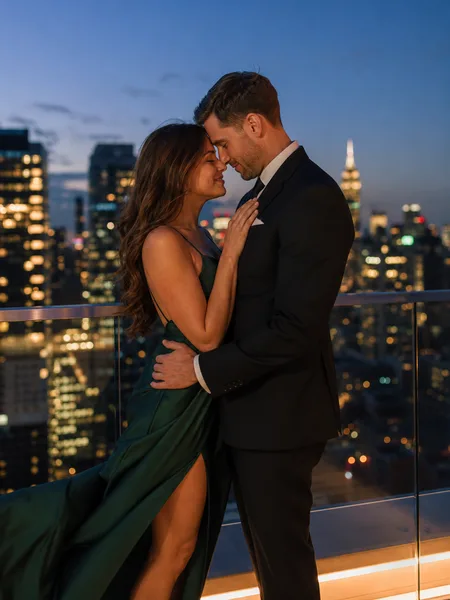 Couple in formal attire touching foreheads on a rooftop at blue hour with city lights behind, engagement photos