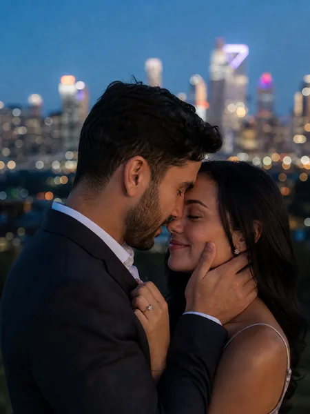 Couple in formal attire touching foreheads at blue hour with city skyline behind them, engagement photos