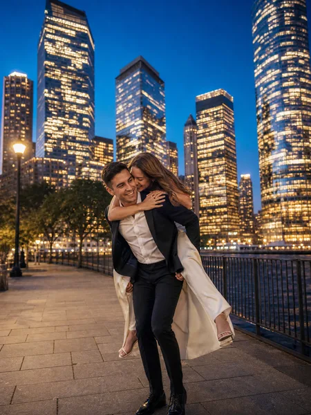 Couple in formal wear with her riding piggyback, city skyline at blue hour behind them, engagement photos