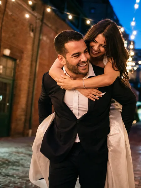 Man carrying woman piggyback style in formal wear, brick alley with string lights, blue hour ambiance, editorial engagement photos