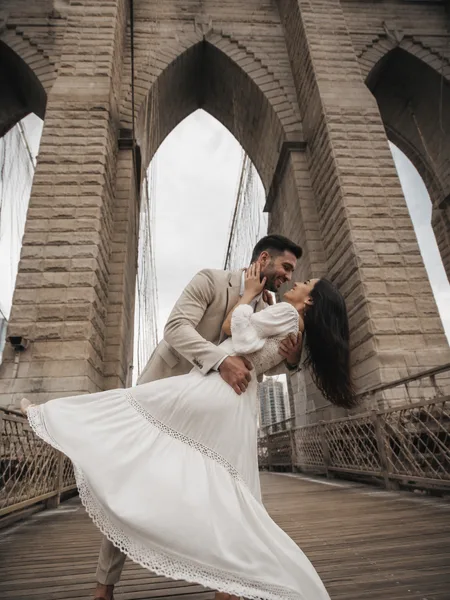 Man dipping woman for a kiss on a historic bridge, white dress flowing, soft cloudy sky, engagement photos