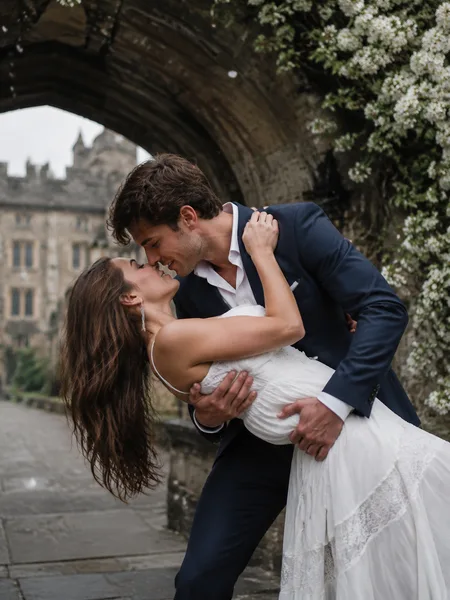 Couple sharing a kiss in a gentle dip under a white blossom-covered archway, editorial engagement photos.