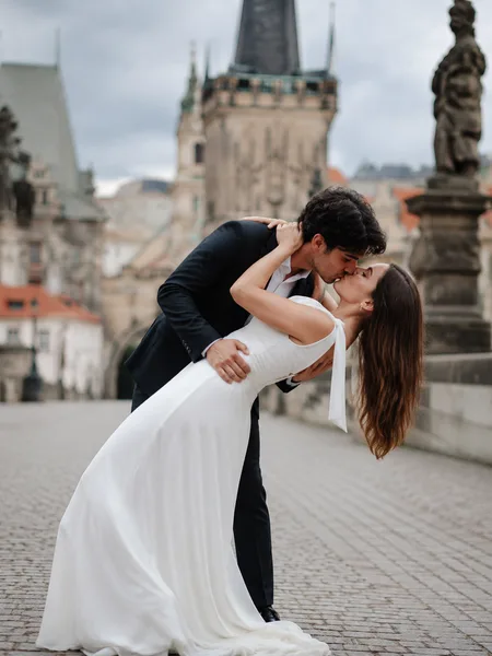 Gentle dip kiss of couple in white dress and suit on stone bridge in soft overcast light, engagement photos.