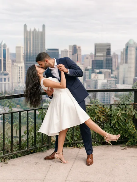 Groom dipping bride in white dress for a kiss on park balcony with city skyline engagement photos