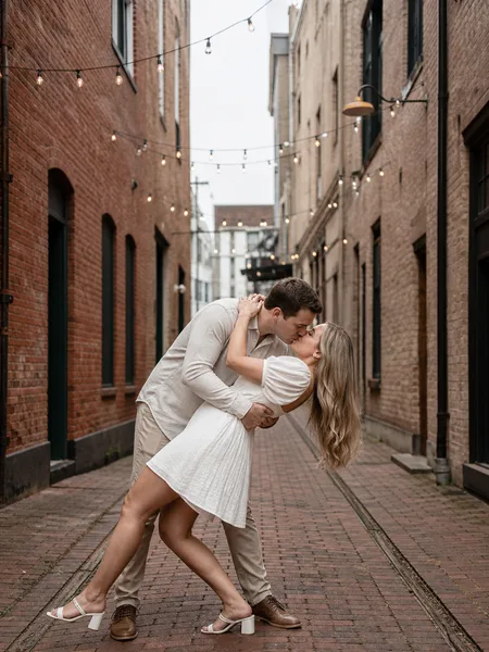 Couple in a gentle dip kiss, woman in white dress, brick alley with string lights, soft daylight, playful engagement photos