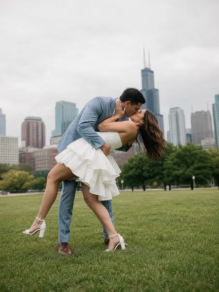 Couple in a gentle dip kiss in a park, woman in white dress, city skyline faint in background, engagement photos