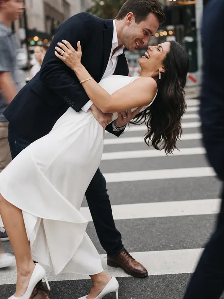 Engagement photos: Playful dip kiss on a crosswalk, bride in white dress laughing, soft overcast light, city backdrop.