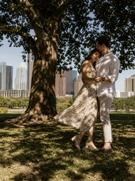 Couple dancing in a leafy park with the city skyline faintly in the distance, engagement photos