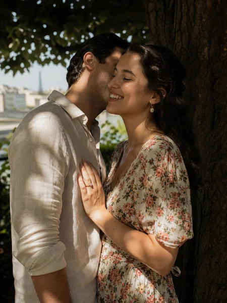 He whispers to her in a shaded park, floral dress, city skyline seen through leaves, engagement photos