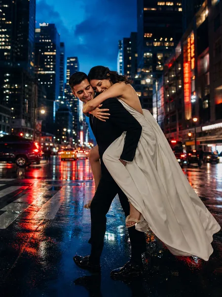 Engagement photos: Groom giving bride a piggyback ride on a busy crosswalk at blue hour, formal attire, city lights reflected on wet street.