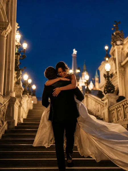 Formal-clad couple playfully piggybacking on a grand staircase with city lights at twilight, engagement photos