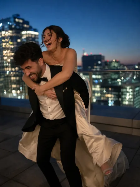 Couple in formal wear piggyback riding on a rooftop at blue hour with city lights, engagement photos