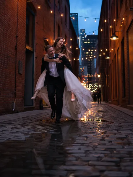 Groom giving bride piggyback ride in brick alley at blue hour, engagement photos.