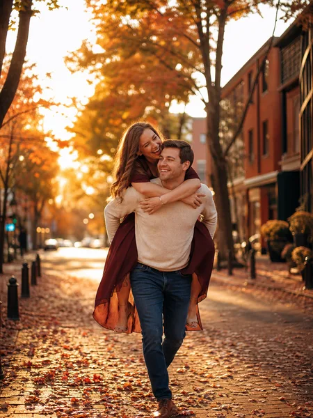 Engagement couple piggyback riding on a leaf-covered city street in golden hour light.