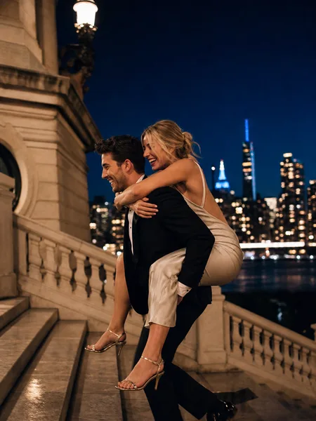 Playful piggyback ride on a grand staircase at twilight with city lights and formal wear, engagement photos