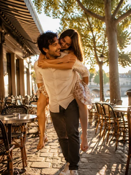A couple shares a piggyback ride between cafe tables on a sun-drenched terrace, her floral dress swaying, golden light streaming through trees, engagement photos.