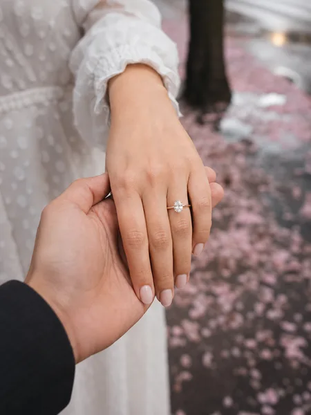 A close-up of a couple’s hands revealing an engagement ring against a backdrop of fallen cherry blossoms on a city street, editorial engagement photos.