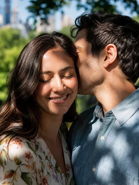Couple in floral dress and chambray shirt, whispering under tree shade with city skyline engagement photos