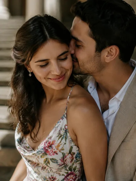 Couple whispering intimately on a shaded grand staircase, woman in floral dress smiling, engagement photos