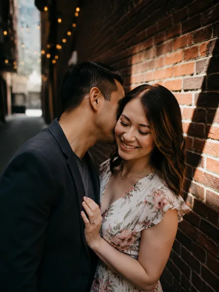 Man whispering to woman in floral dress, brick alley with string lights, dramatic midday shade, intimate engagement photos