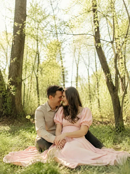 A couple sitting closely on grass in a spring forest with new green leaves and soft morning sunlight, engagement photos.