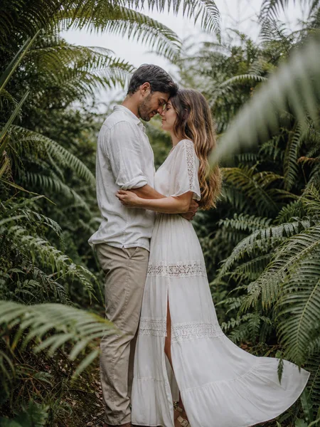 A couple stands with foreheads touching on a fern-lined forest trail, her boho dress flowing, engagement photos.