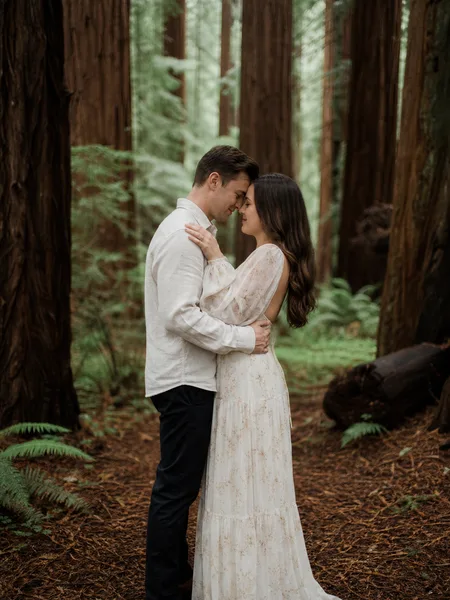 Couple with foreheads touching, standing in a redwood forest with overcast light, for engagement photos.