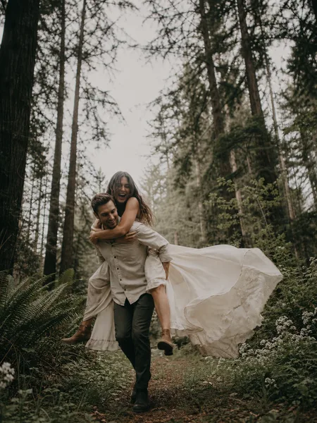 Piggyback ride in a forest clearing, woman in flowy boho dress, soft overcast light, engagement photos.
