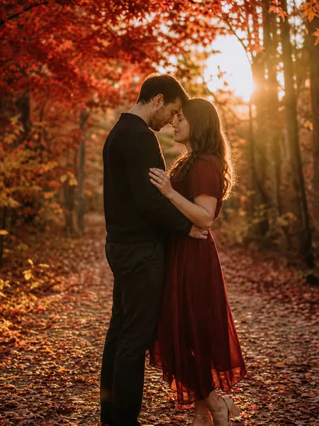 Couple with foreheads touching, standing on a forest path blanketed with red and orange maple leaves, bathed in golden hour sunlight during engagement photos.