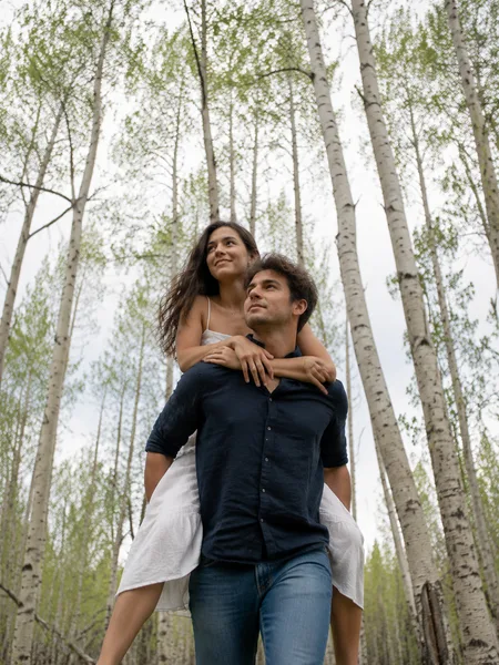 A woman rides piggyback on a man in a birch forest, white dress, navy shirt, engagement photos