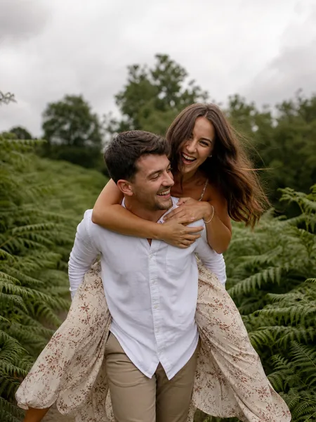 A groom gives a bride a piggyback ride on a fern-lined forest trail, her boho dress trailing, engagement photos.