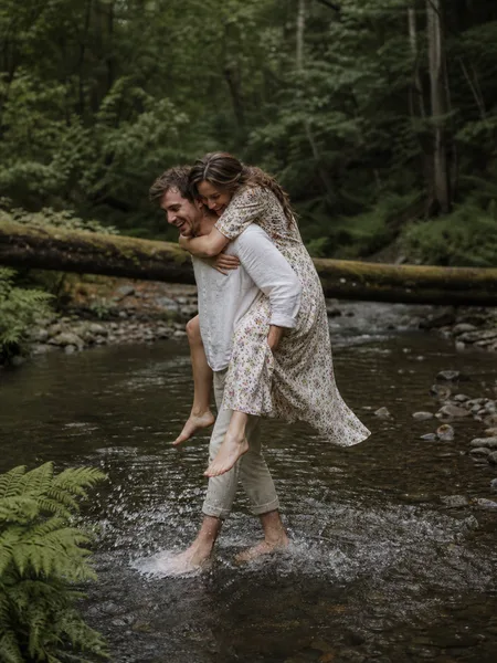 Wide shot of couple with piggyback ride by mossy log and stream, flowy boho dress, overcast light, dreamy engagement photos