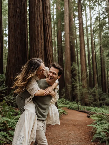 A man giving a woman a piggyback ride in a redwood forest, with soft overcast light, for engagement photos.