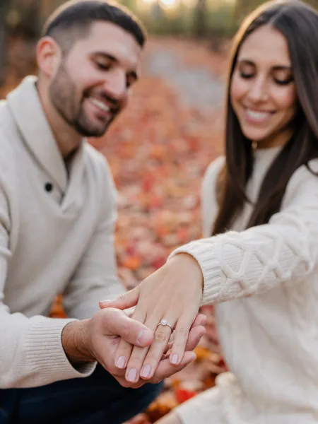 Close-up of a couple's hands with an engagement ring revealed, kneeling on red and orange maple leaves, soft afternoon light, during engagement photos.