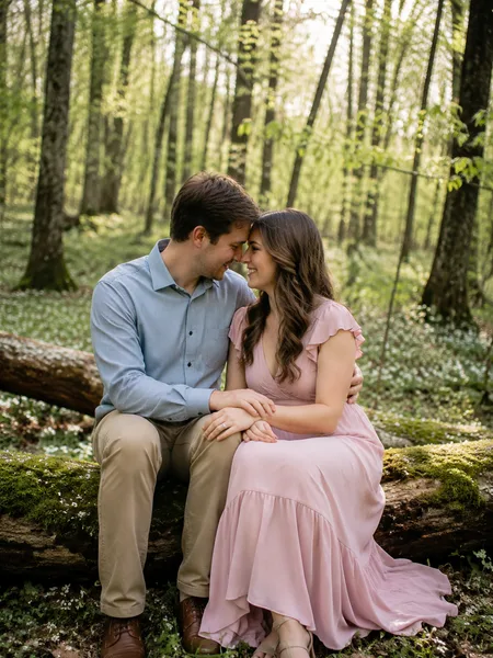 A couple sits close on a mossy log in a spring forest, wearing a pink dress and chambray shirt, engagement photos
