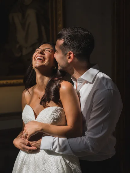 Couple in white sharing a candid laugh, dramatic side light creating a chiaroscuro effect in a gallery, engagement photos.